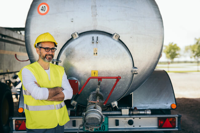 Trabalhador da construção civil de braços cruzados e vestindo EPIs, posando atrás de caminhão pipa.